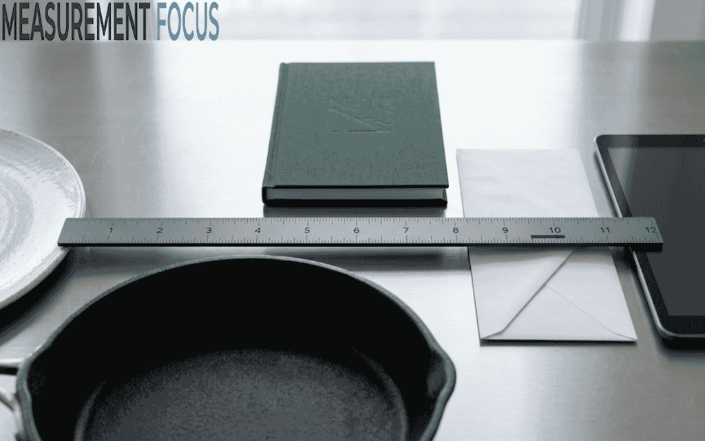 A ruler rests on a surface alongside a book, envelope, tablet, plate, and skillet
