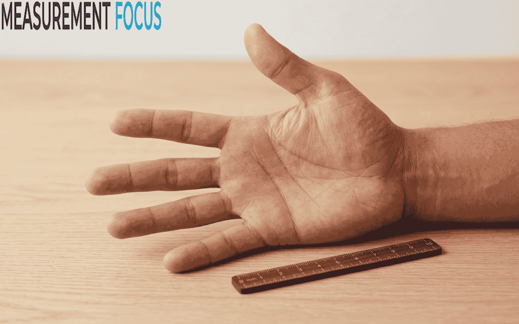 An adult's open right hand with palm facing up rests on a wooden table, next to a 10-centimeter-long wooden ruler.