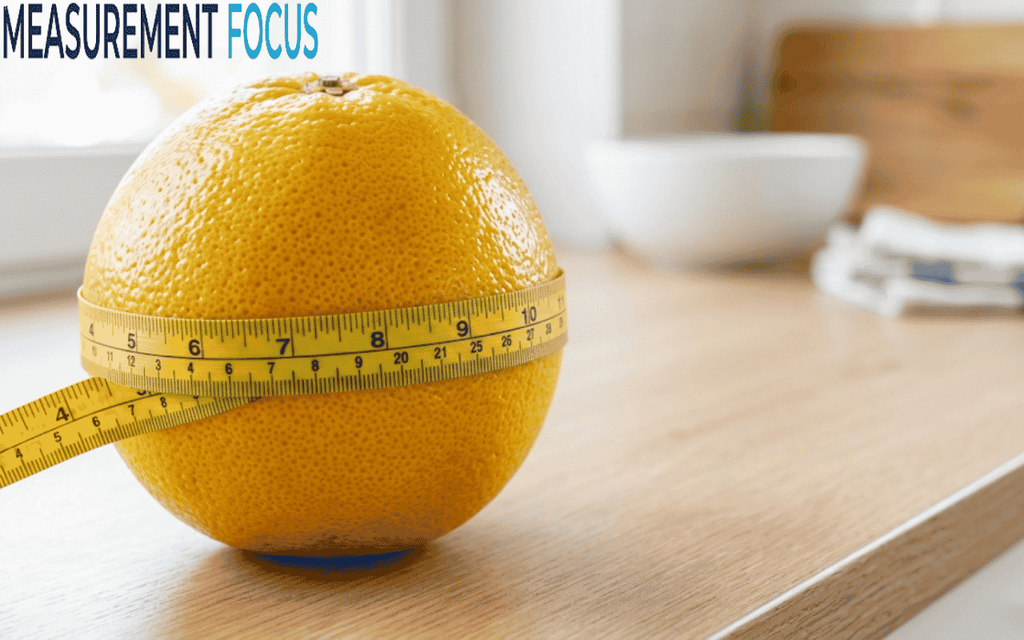 An orange on a wooden counter is wrapped with a yellow measuring tape, which reads from 4 to 10 inches. Background objects are blurred.