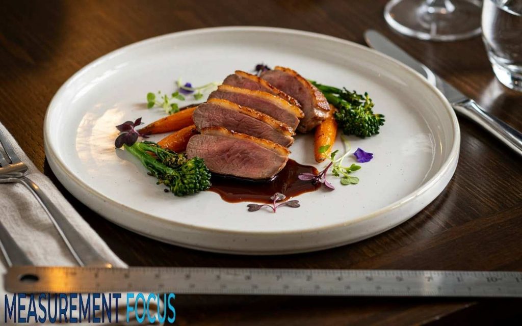 Sliced medium rare duck breast with roasted carrots and broccolini on a white plate next to a metal ruler on a wooden table.